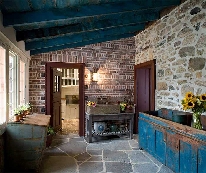 photo of a sunroom in an 1829 house one wall is brick. One wall is fieldstone. One wall is windows. Exposed beams and wood ceiling that seem to be stained blue. There is a flagstone or slate floor. An old dry sink sits along the fieldstone wall. Addition by Peter Zimmerman Architects and shown on Town N Country Living via the happy list