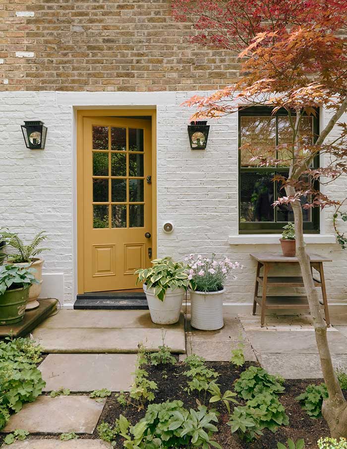 brick house painted white with a yellow front door. The door has a grid of 12 windows. There are two black lanterns on either side of the front door. There's a flagstone path leading to the front door with concrete planters filled with green plants. Photo taken by Michael sinclair for House and Garden UK and featured on the Happy List.
