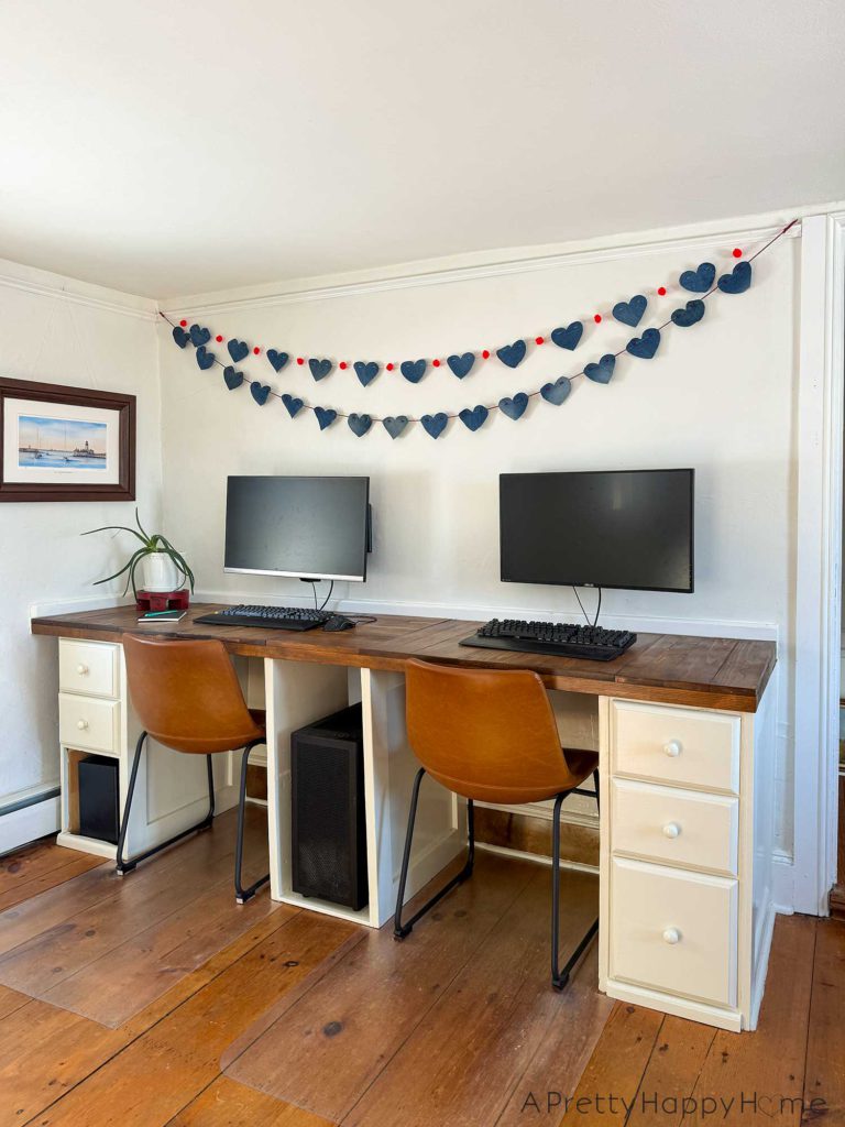 a no sew denim heart bunting banner hanging above a monitor on a wood desk. The hearts on the bunting are made from denim and are 4 inches wide. They are hung on a red cord. There's a second denim heart bunting that has red pom poms in between each heart and hanging on the wall.