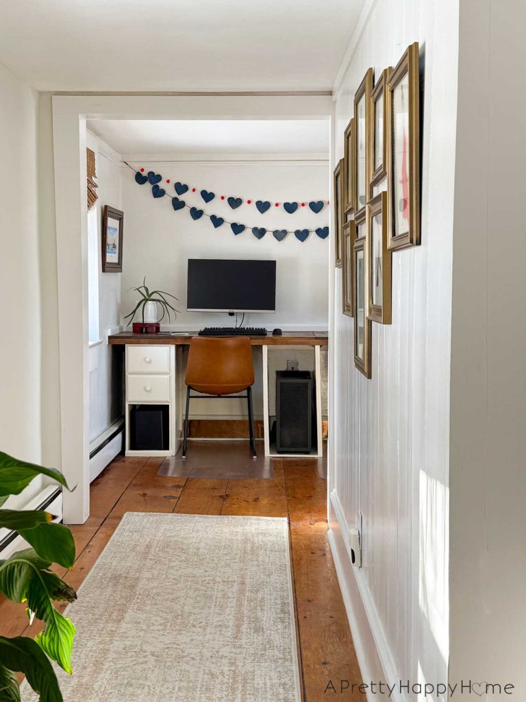 a no sew denim heart bunting banner hanging above a monitor on a wood desk. The hearts on the bunting are made from denim and are 4 inches wide. They are hung on a red cord. There's a second denim heart bunting that has red pom poms in between each heart and hanging on the wall. In the foreground of the picture is a hallway with a neutral rug runner and gold picture frames on the wall.