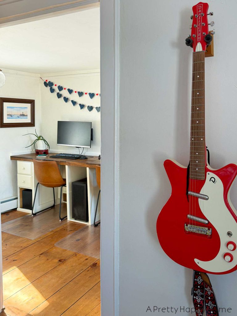 a no sew denim heart bunting banner hanging above a monitor on a wood desk. The hearts on the bunting are made from denim and are 4 inches wide. They are hung on a red cord. There's a second denim heart bunting that has red pom poms in between each heart and hanging on the wall. In the foreground of the picture is a red and white electric guitar.