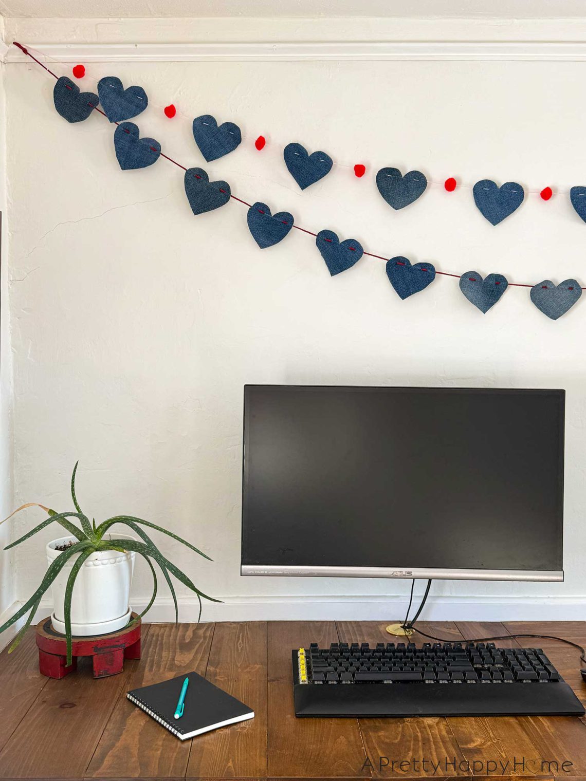 a no sew denim heart bunting banner hanging above a monitor on a wood desk. The hearts on the bunting are made from denim and are 4 inches wide. They are hung on a red cord. There's a second denim heart bunting that has red pom poms in between each heart and hanging on the wall.