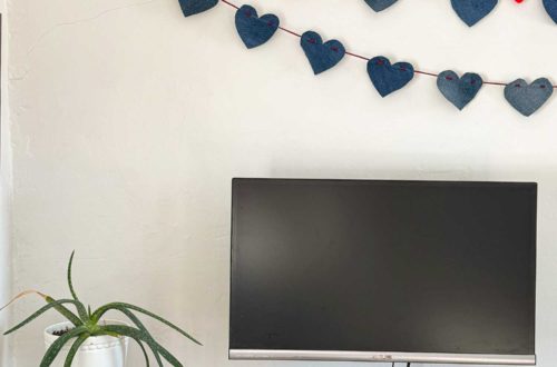 a no sew denim heart bunting banner hanging above a monitor on a wood desk. The hearts on the bunting are made from denim and are 4 inches wide. They are hung on a red cord. There's a second denim heart bunting that has red pom poms in between each heart and hanging on the wall.