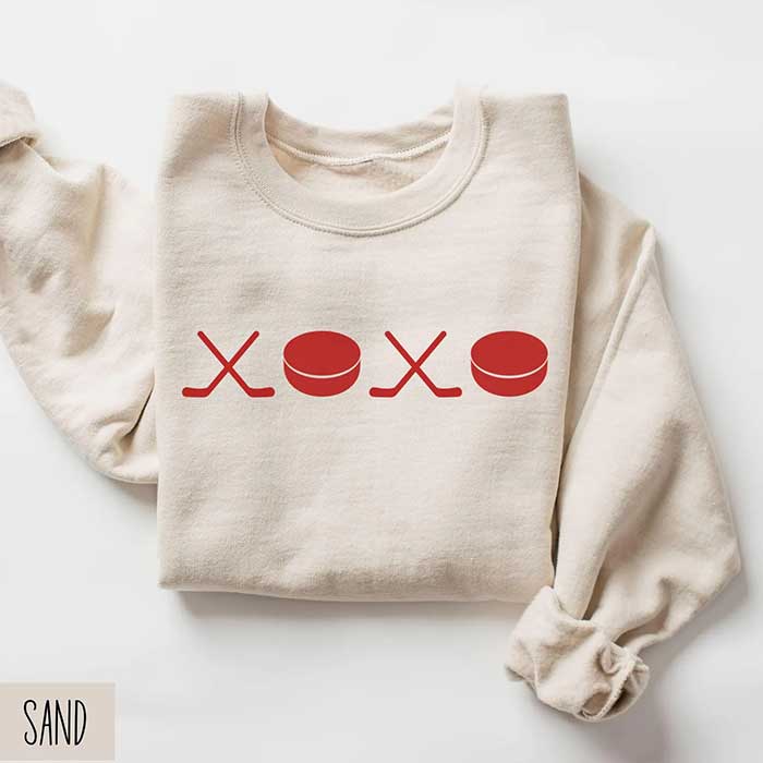 sand colored sweatshirt showing xoxo but the x's are made from hockey sticks and the o's are pucks. It's a hockey valentine sweatshirt made by etsy shop watermelonhoneyco and shown here on the happy list