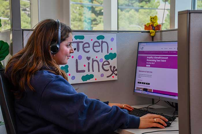 photo of a teen sitting at a computer cubicle volunteering for Teen Line, a mental health crisis hotline for teens. Photo courtesy of Teen Line and found at Reasons to be Cheerful.