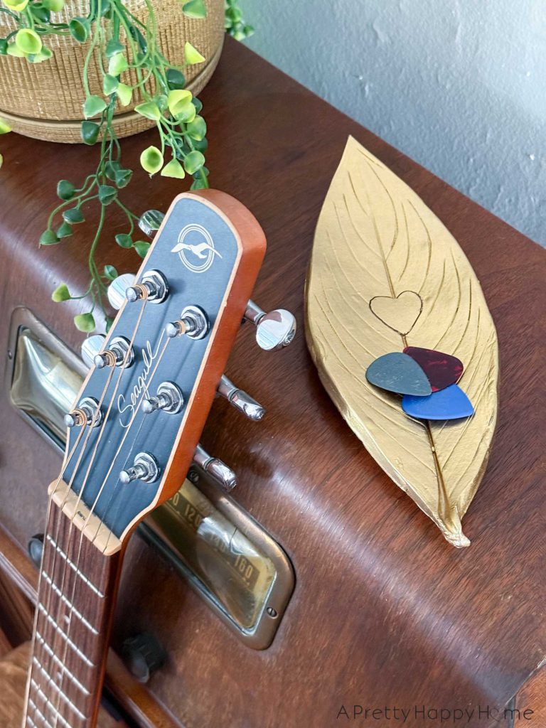 photo of a DIY Polymer Clay Leaf Dish with Heart Detail. The small trinket dish is painted gold and is holding guitar picks because there's a guitar in the foreground of the photo. The heart detail in the center of the trinket dish was achieved by cutting a heart shape out of the leaf prior to pressing the leaf into the clay to make an impression.