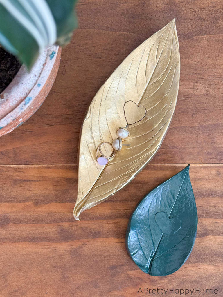 photo of two DIY Polymer Clay Leaf Dishs with Heart Detail. One of the small trinket dishes is painted gold, the other is green. The gold dish is holding a pair of pearl earrings an a ring with a pink stone. The heart detail in the center of the trinket dish was achieved by cutting a heart shape out of the leaf prior to pressing the leaf into the clay to make an impression.