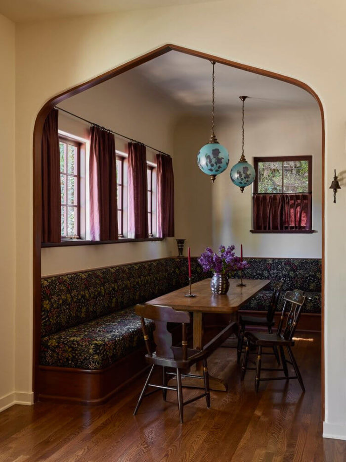 dining room in a 1906 tudor home featuring a built in banquette on two walls with a narrow dining table in front of it. There are 4 windows above the banquette with burgundy curtains. Midcentury style turquoise globe pendants hang above the table. The entire room is rich yet inviting. Design by Reath Design and photo by Laure Joliet. Featured on Desire to Inspire.