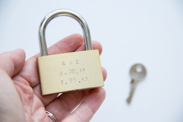 photo of a hand holding diy anniversary lock with the initials of the couple and their wedding anniversary date stamped on it with a metal stamping kit. Idea from Lovely Indeed.