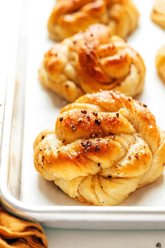 kardemummabullar or Swedish Cardamom Rolls sitting on a baking sheet. The rolls are golden brown with a twisted dough look, and have a sprinkle of cardamom on top. Recipe by Gimme Some Oven and shown here on the happy list.