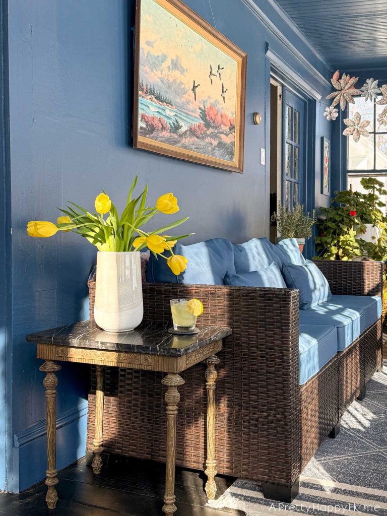 ornate vintage brass and soapstone table sitting next to a rattan couch with blue cushions in a sunporch painted navy blue with a painted black wood floor. There is a large landscape painting behind the couch. On the table are a vase with yellow tulips and a glass of lemonade.