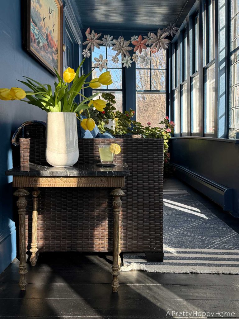 ornate vintage brass and soapstone table sitting next to a rattan couch in a sunporch painted navy blue with a painted black wood floor. There are a set of stained glass doors behind the table. on the table are a vase with yellow tulips and a glass of lemonade.