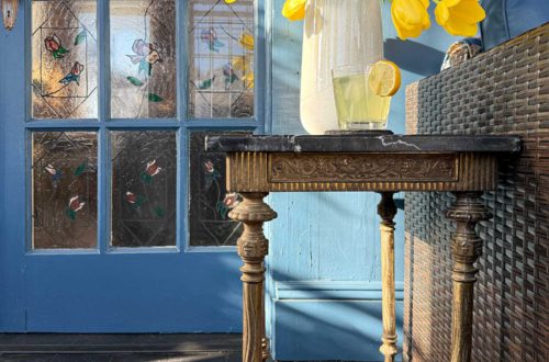 ornate vintage brass and soapstone table sitting next to a rattan couch in a sunporch painted navy blue with a painted black wood floor. There are a set of stained glass doors behind the table. on the table are a vase with yellow tulips and a glass of lemonade.