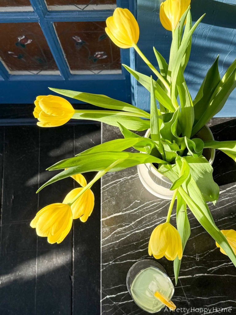 ornate vintage brass and soapstone table sitting next to a rattan couch in a sunporch painted navy blue with a painted black wood floor. There are a set of stained glass doors behind the table. on the table are a vase with yellow tulips and a glass of lemonade.
