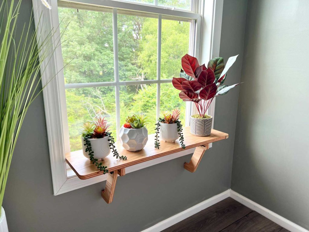 photo of an oak window plant ledge created by etsy shop TheNamu. It sits on a white windowsill with grey walls. There are four small plants sitting on the ledge.Included in a blog post about unique plant displays.