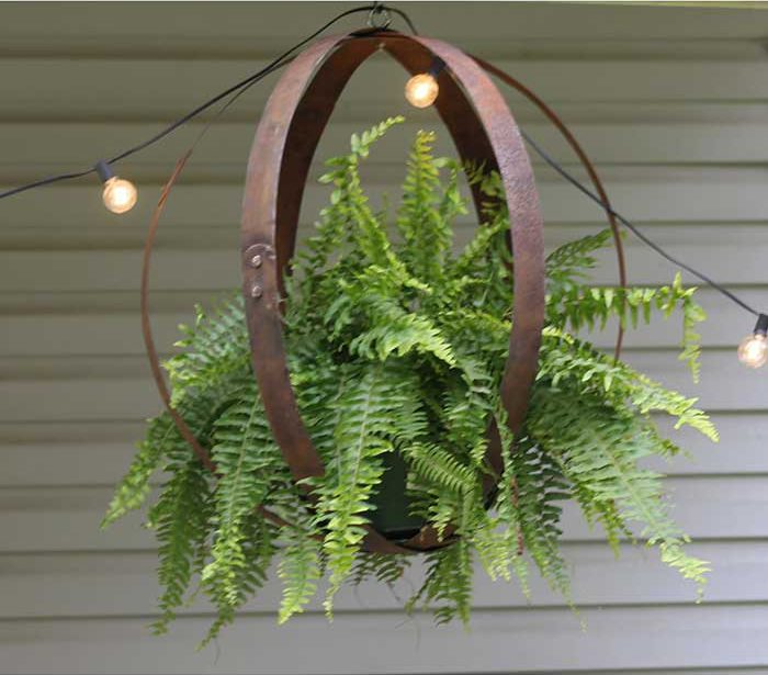 photo of a barrel ring hanging plant holder that's holding a fern. The barrel rings form an orb around the fern. There are string lights hung above the plant as it seems to be on a front porch. Sold by Walmart. Shown here on unique plant display ideas blog post.