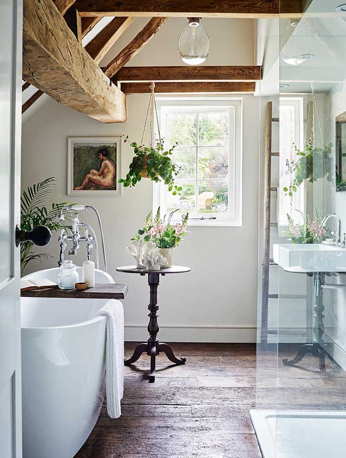 rustic bathroom with wood beams, wood floor, freestanding soaking tub, pedestal table with flowers, a window with a garden view and a glass enclosed shower. photo by brent darby via homes and gardens,