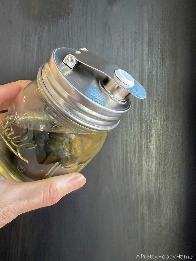 mason jar containing simple syrup tip slightly to showcase a lid with a flip top and pour spout. It's made with stainless steel and silicone. No extra plastic. Sold on Amazon.