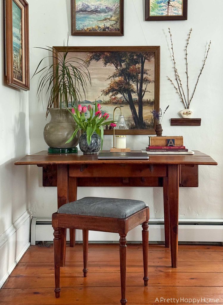 wood stool with a grey leather upholstered top sitting in front of a rustic drop leaf table. On the table is a plant, a charcoal vase with pink tulips, a laptop, a candle warmer lamp, and a stack of books. Behind the table is a gallery wall of nature paintings.