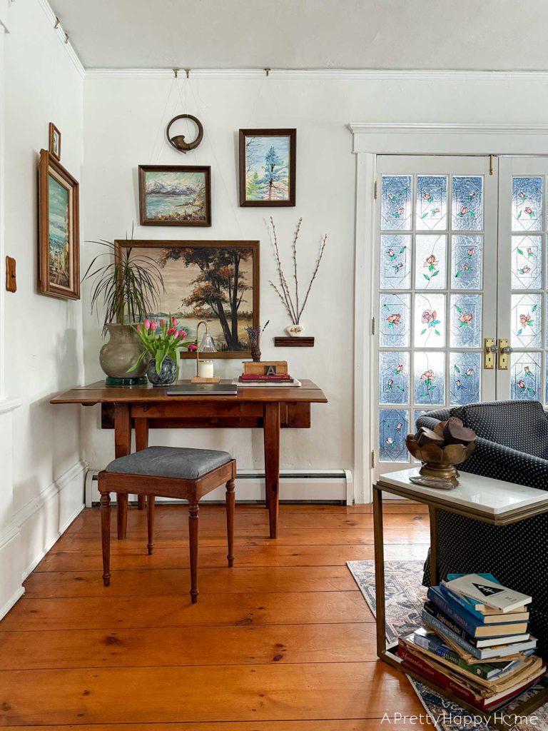 wood stool with a grey leather upholstered top sitting in front of a rustic drop leaf table. On the table is a plant, a charcoal vase with pink tulips, a laptop, a candle warmer lamp, and a stack of books. Behind the table is a gallery wall of nature paintings.There are french doors with stained glass to the right of the table.