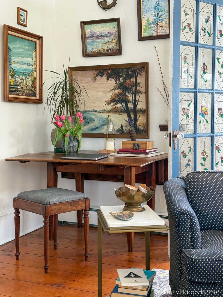 wood stool with a grey leather upholstered top sitting in front of a rustic drop leaf table. On the table is a plant, a charcoal vase with pink tulips, a laptop, a candle warmer lamp, and a stack of books. Behind the table is a gallery wall of nature paintings.There is a blue french door with stained glass to the right of the table.