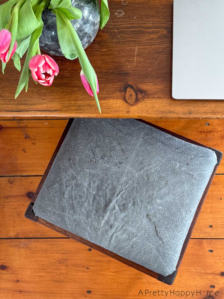 wood stool with a grey leather upholstered top sitting in front of a rustic drop leaf table. On the table is a charcoal vase with pink tulips and a laptop.