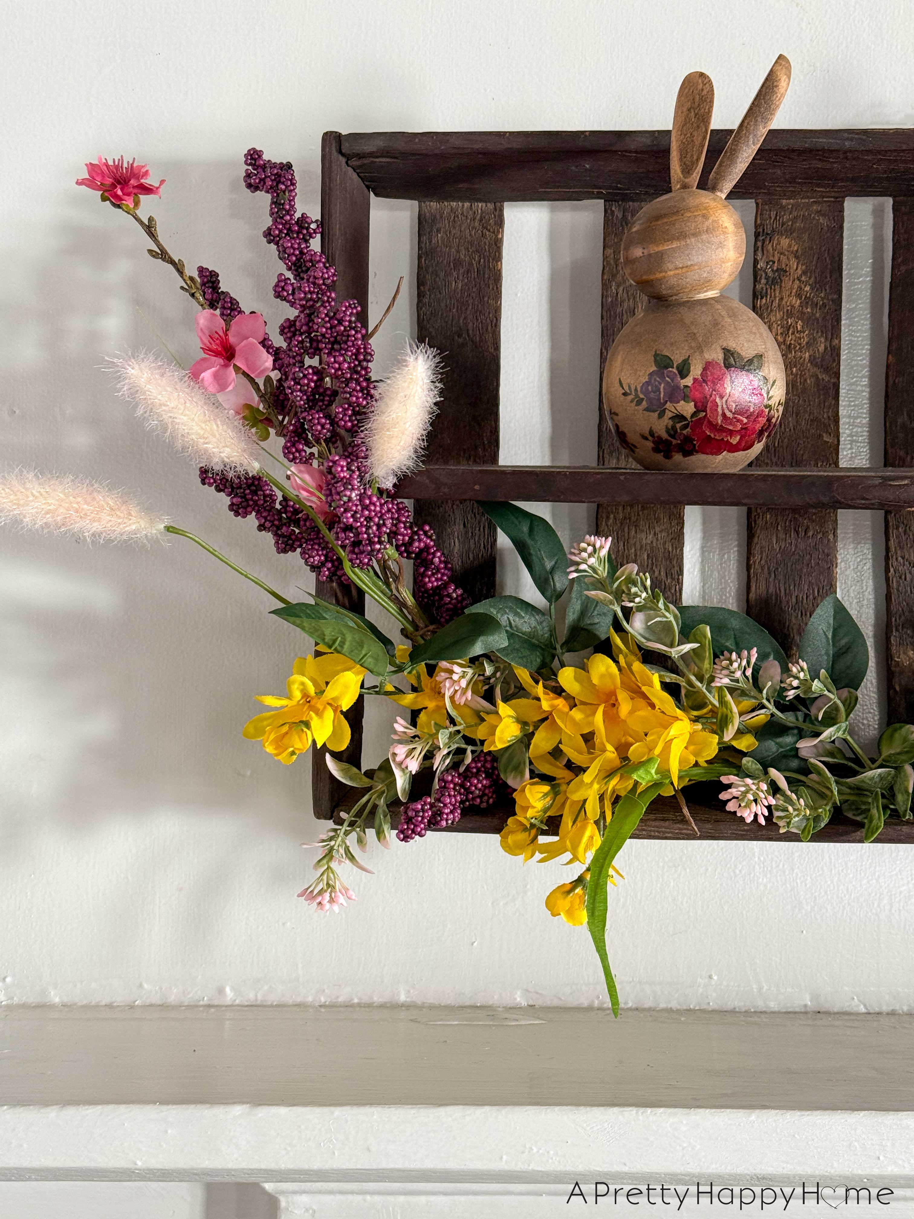 diy repurposed berry crate spring wreath using a vintage wood berry field flat or berry basket carrier. It's hanging on the wall with sprays of pink, yellow, and purple faux flowers attached to it. There's a wood bunny decorated with painted flowers sitting on the wreath.
