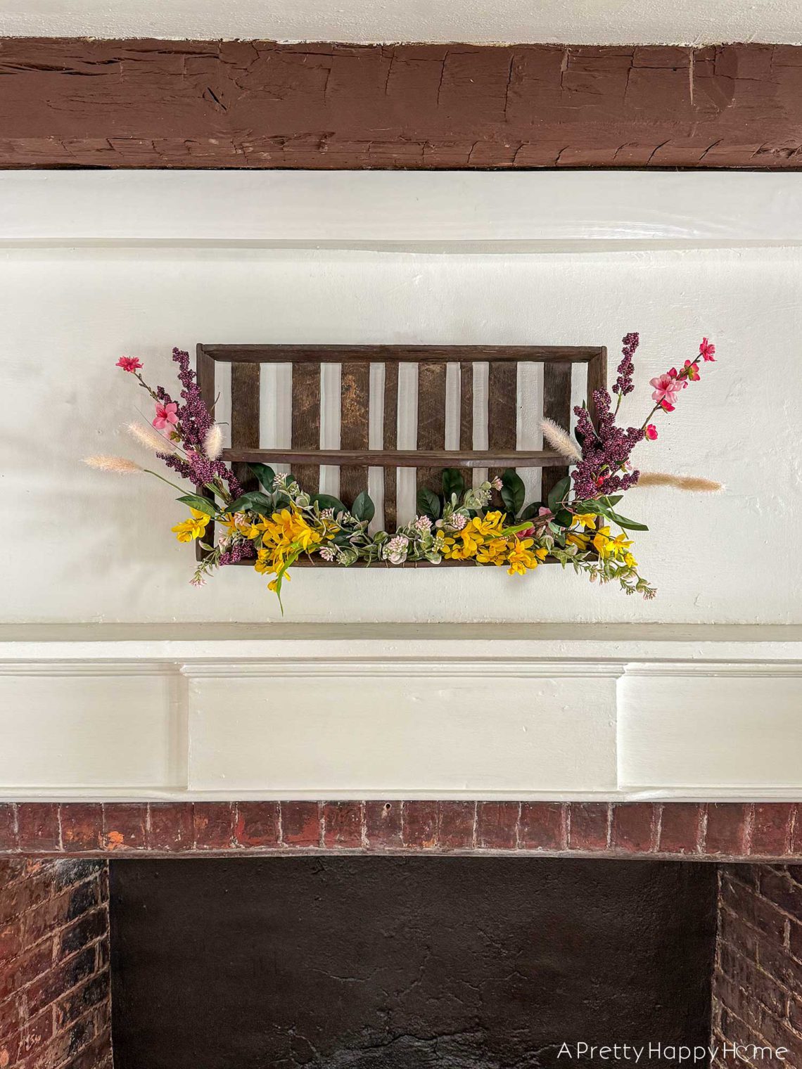 diy repurposed berry crate spring wreath using a vintage wood berry field flat or berry basket carrier. It's hanging on the wall with sprays of pink, yellow, and purple faux flowers attached to it.