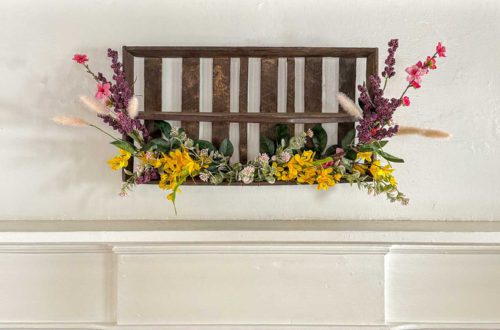 diy repurposed berry crate spring wreath using a vintage wood berry field flat or berry basket carrier. It's hanging on the wall with sprays of pink, yellow, and purple faux flowers attached to it.