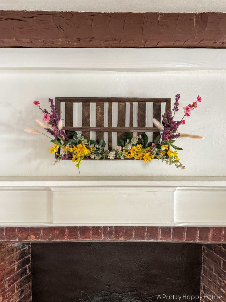 diy repurposed berry crate spring wreath using a vintage wood berry field flat or berry basket carrier. It's hanging on the wall with sprays of pink, yellow, and purple faux flowers attached to it.