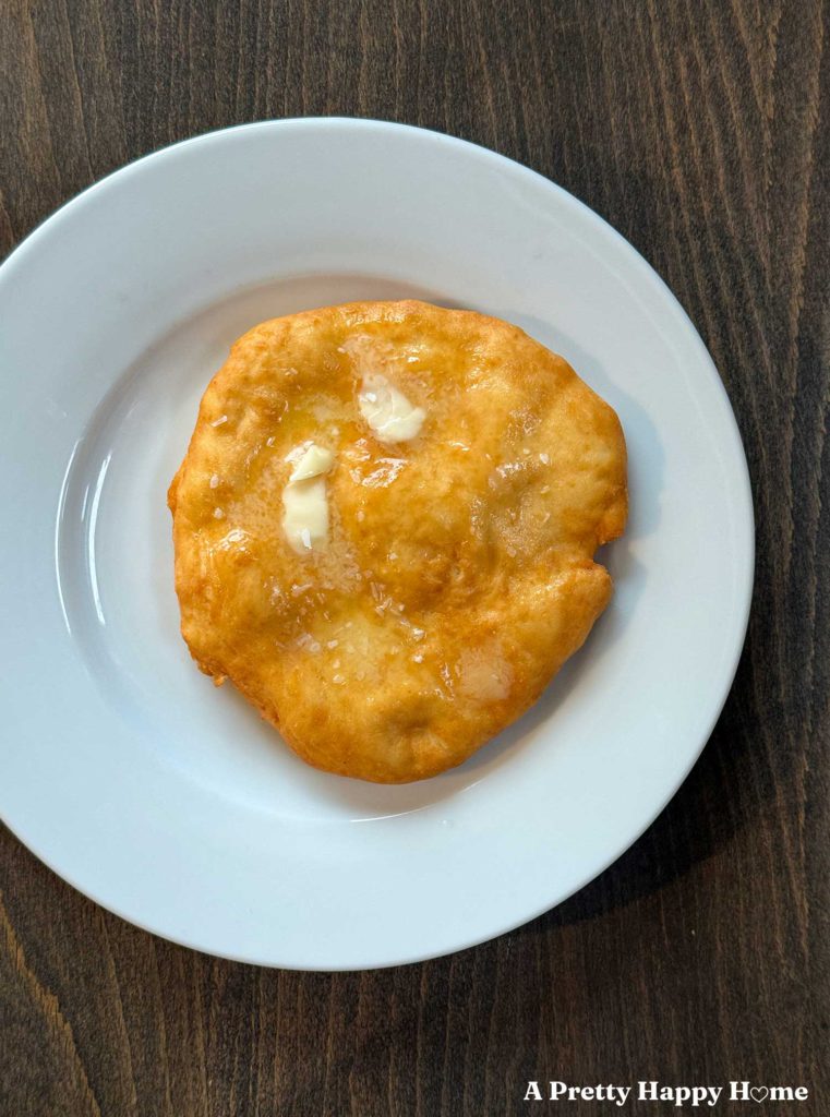 a piece of fry bread, dough fried in oil, sitting on a white plate against a wood background. The fry bread is slathered with butter.