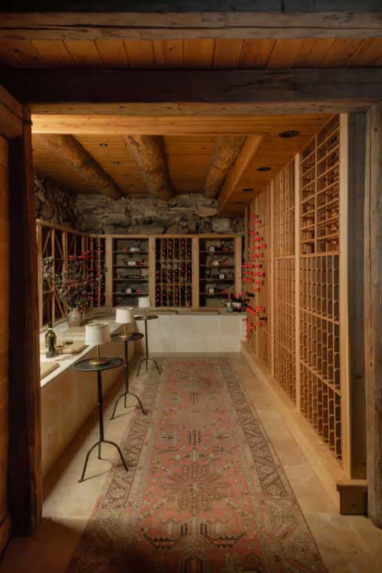 wine cellar situated in a basement with wood beams and stone walls. There's a bench for seating on the left side with 3 small drink tables with tiny lamps on each table. Photo by Carolina Andrade for Style at Home