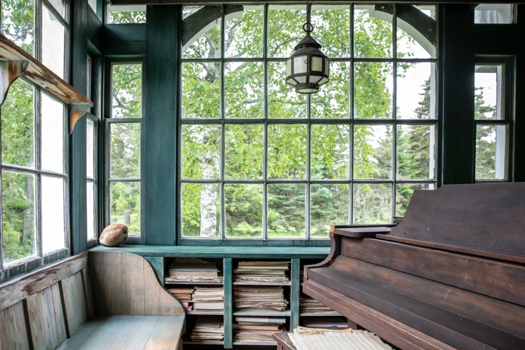 windowed room with green stain on the wood trim, an old grand piano in front of the windows and under the windows is a rustic bench and shelves for sheet music. photo by maura mcevoy for house and garden uk