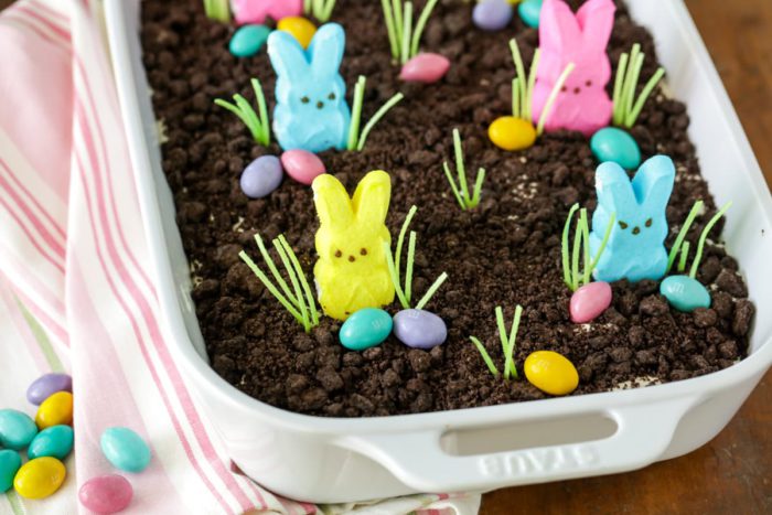 Easter dirt cake in a white casserole dish that is topped with crushed oreo cookies, bunny peeps candy, candy grass, and chocolate easter eggs. recipe by lil luna.