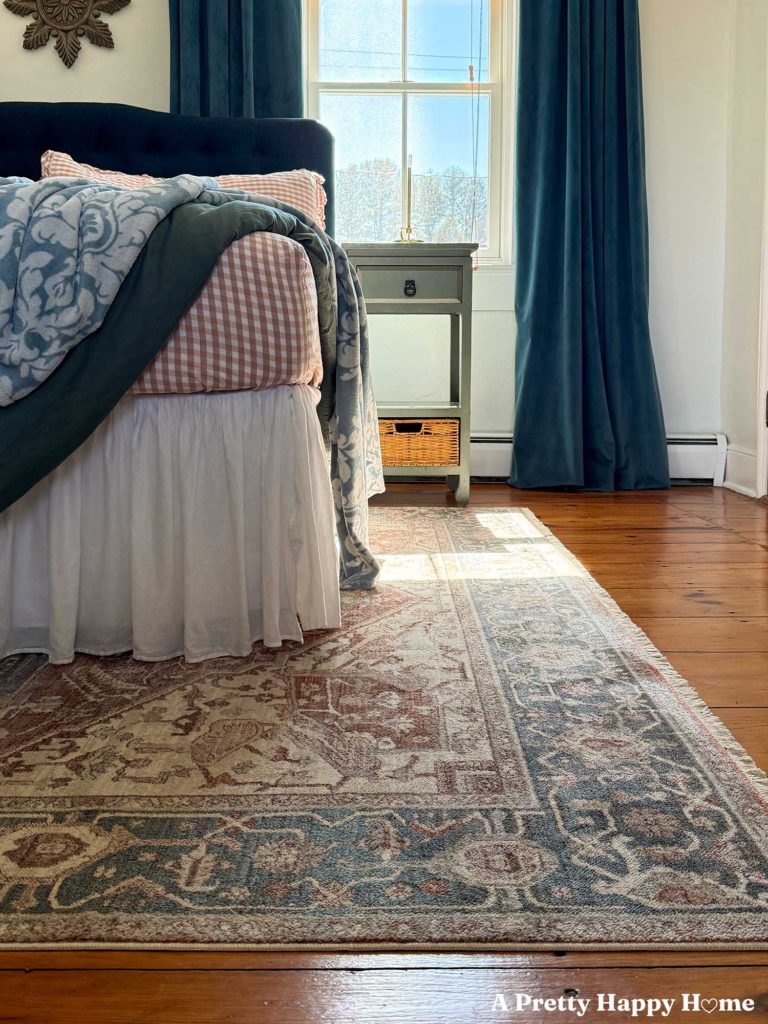 corner of a bed with pink gingham sheets, green duvet, and blue print blanket with a blue and orange rug underneath the bed