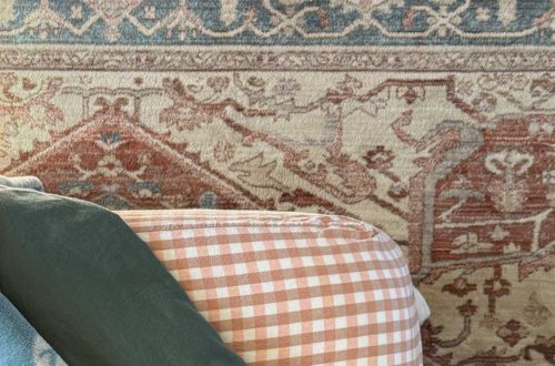 corner of a bed with pink gingham sheets, green duvet, and blue print blanket with a blue and orange rug underneath the bed