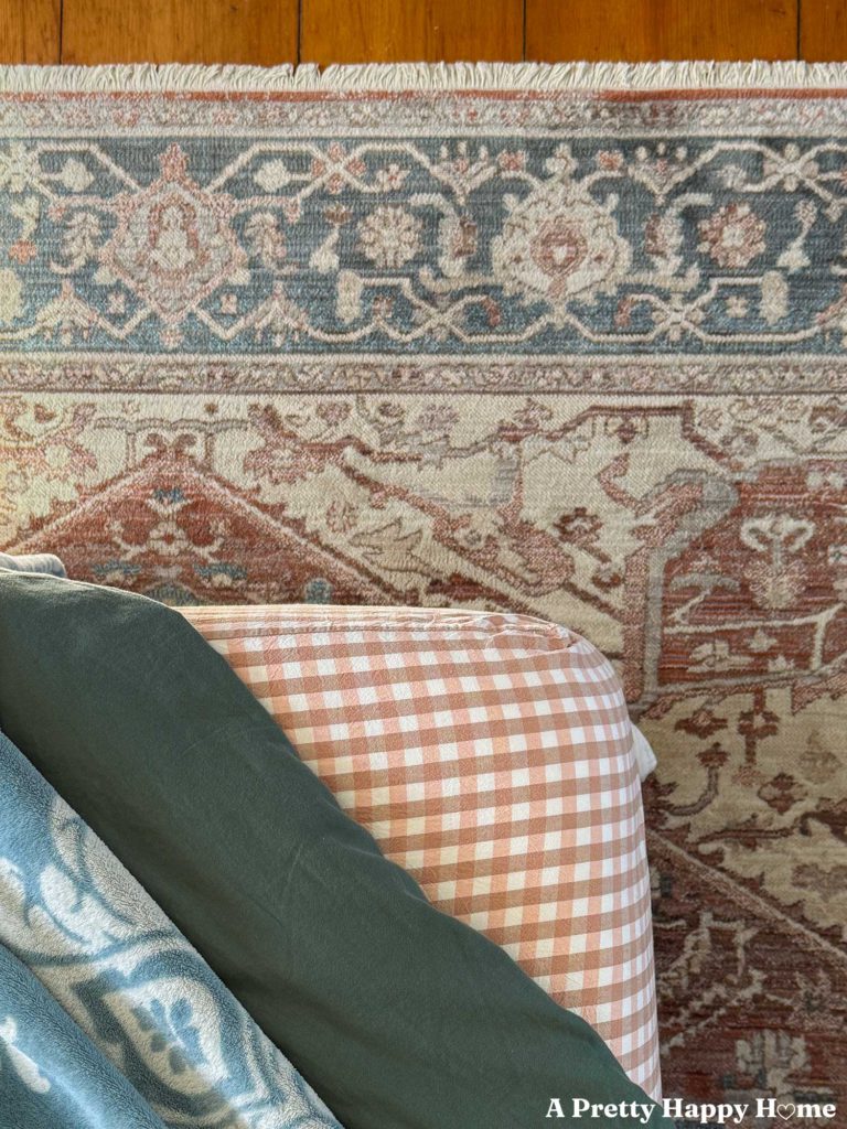 corner of a bed with pink gingham sheets, green duvet, and blue print blanket with a blue and orange rug underneath the bed