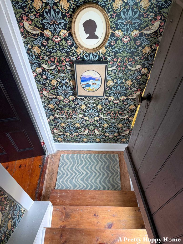 colorful william-morris style wallpaper in a tiny stairwell with wood stairs. There is a striped green rug on the floor and silhouette art and embroidered seagull art on the wall.