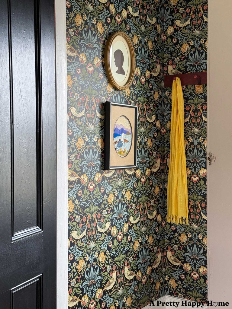 black door opening to colorful william-morris style wallpaper in a tiny stairwell with wood stairs. There silhouette art and embroidered seagull art on the wall hanging next to a red peg rail with a yellow scarf.