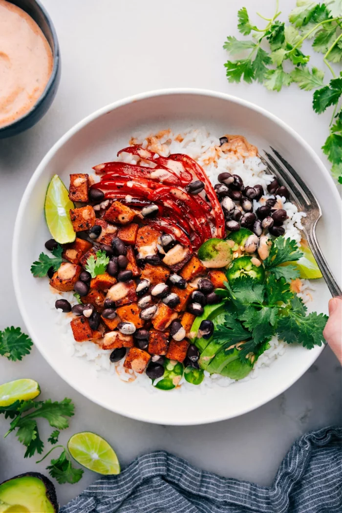 sweet potato and black bean bowl with red peppers, avocado, lime wedges, and cilantro in a white bowl. the recipe is from chelsea's messy apron