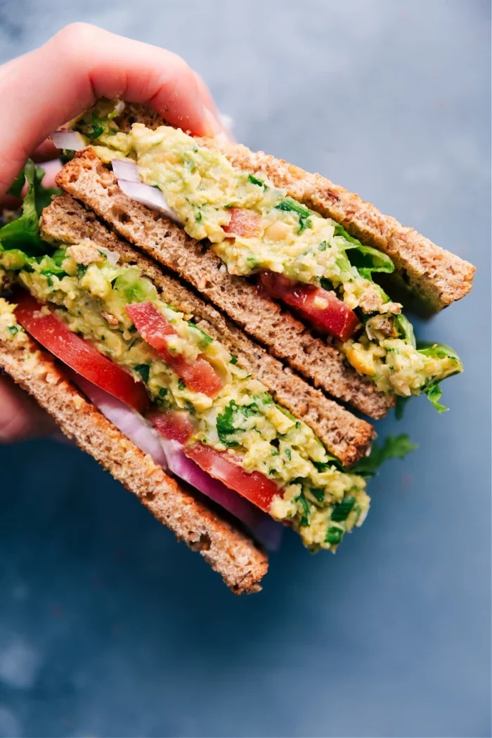 a woman's hand holding a sliced chickpea salad sandwich on toasted bread and stuffed with herbs, tomatoes, and onions. recipe from chelsea's messy apron