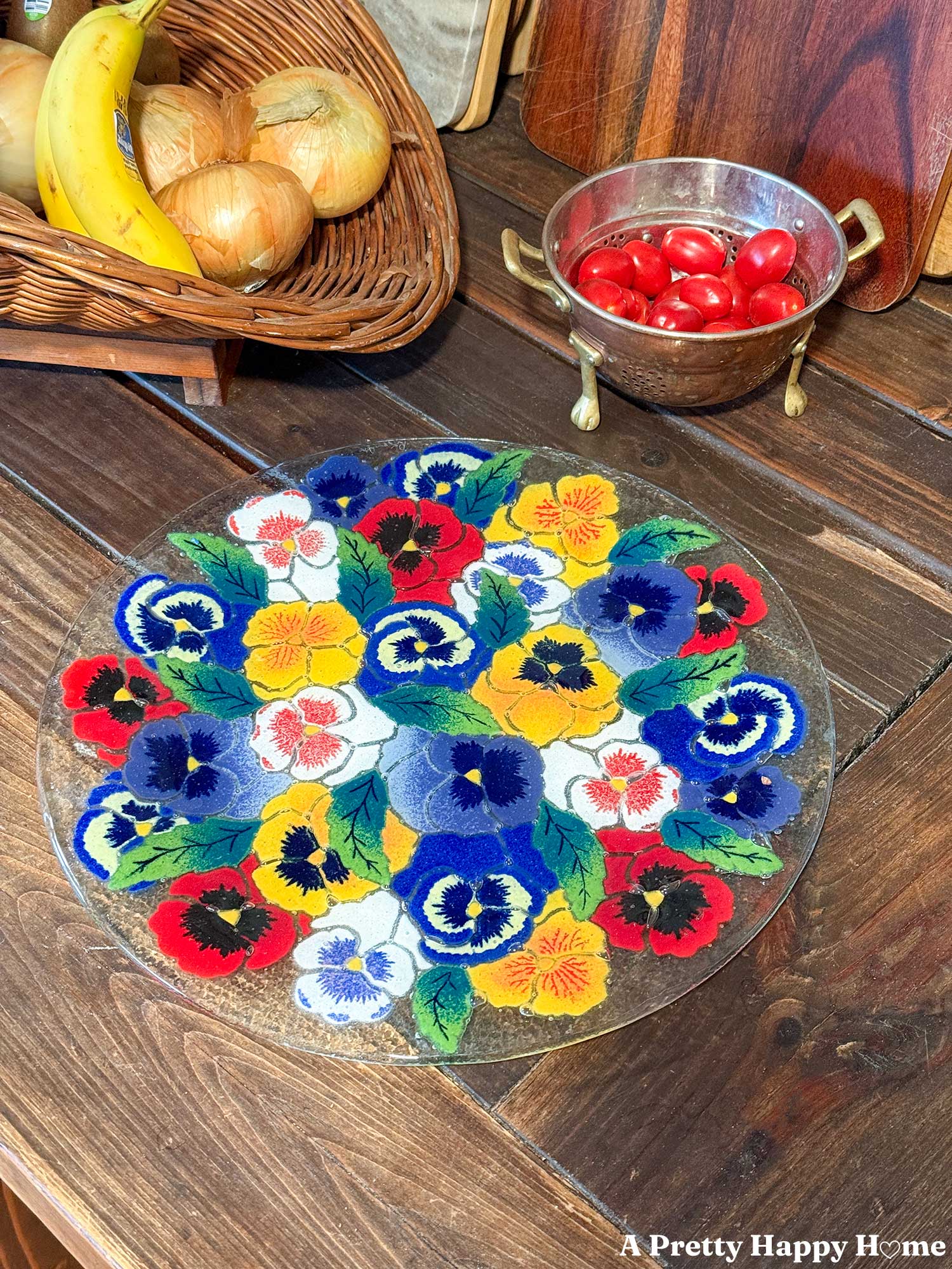a Peggy Karr pansy glass platter sits on a rustic wood table next to a copper colander filled with baby tomatoes and a basket with onions and bananas