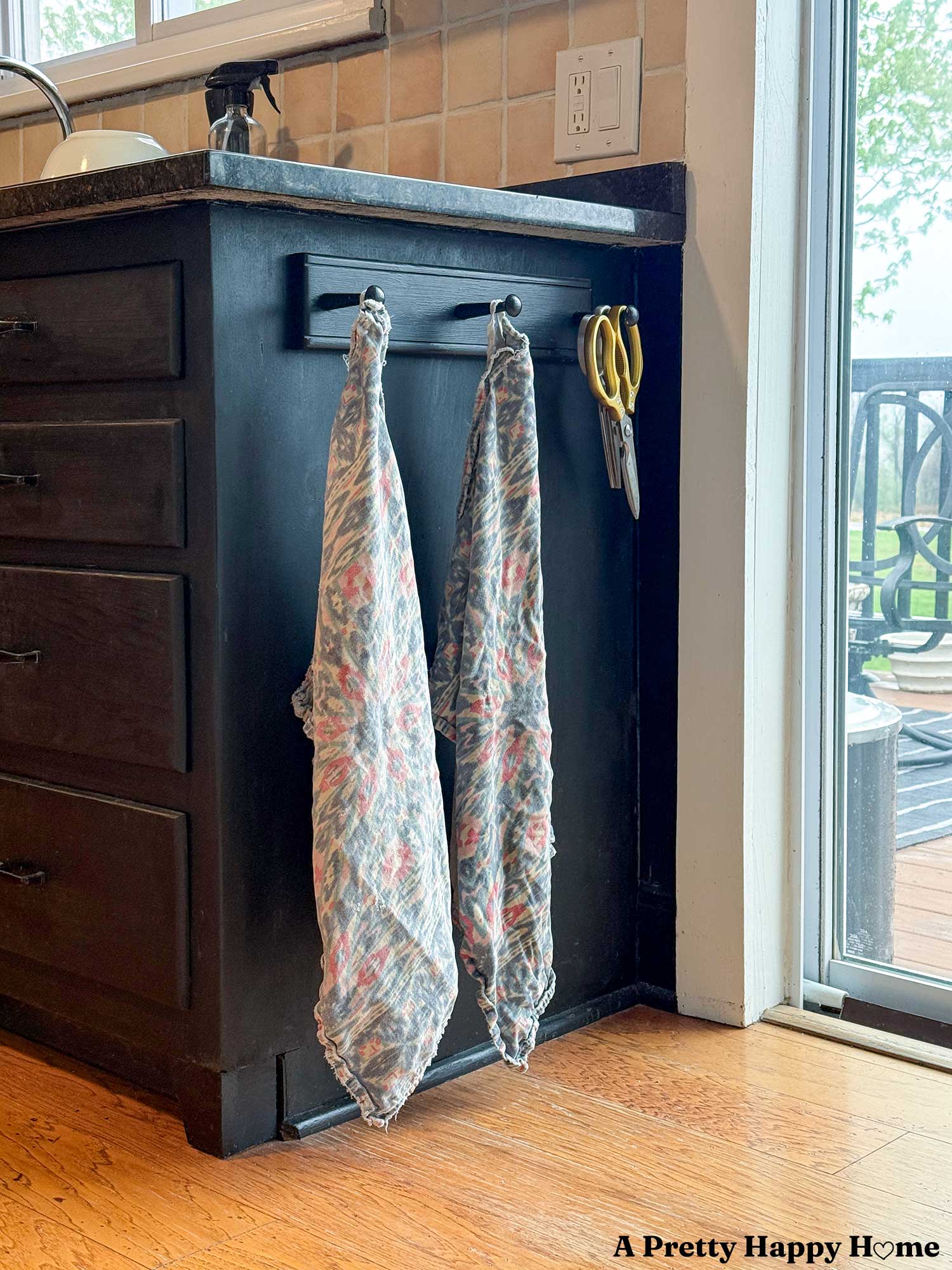 two blue and pink patterened tea towels hang on the end of a black kitchen cabinet from a black peg rail. There is a pair of gold-handed scissors hanging next to the towels.