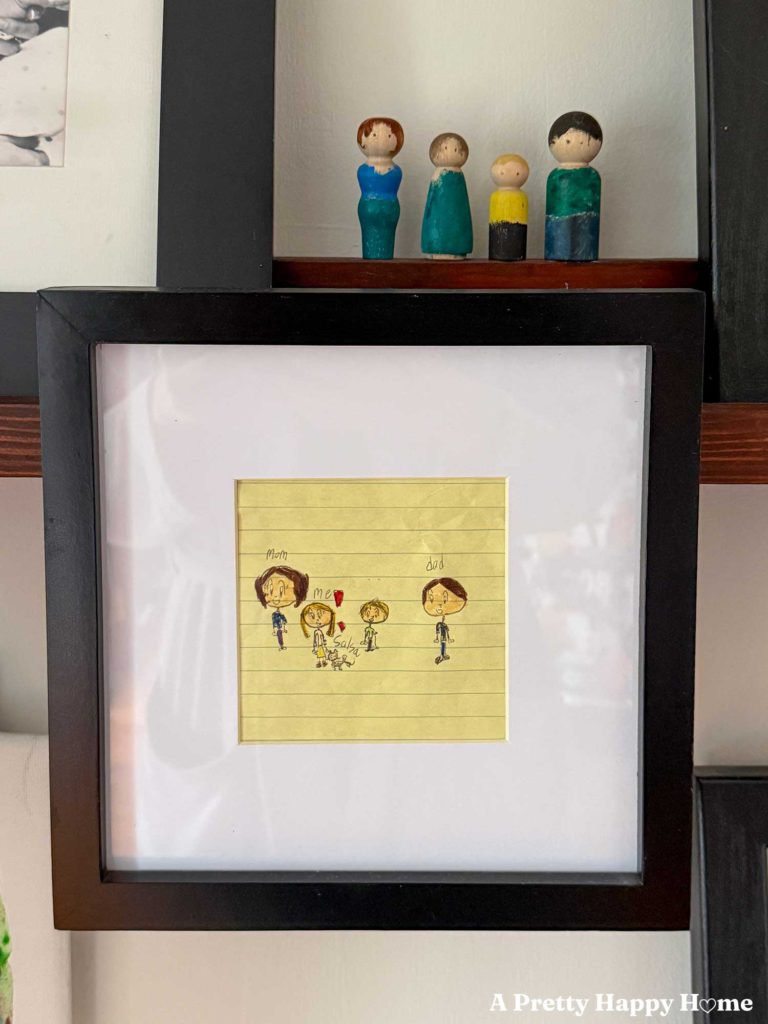 family portrait framed and drawn by a child on yellow lined paper. Above the picture is the same family painted on peg dolls.