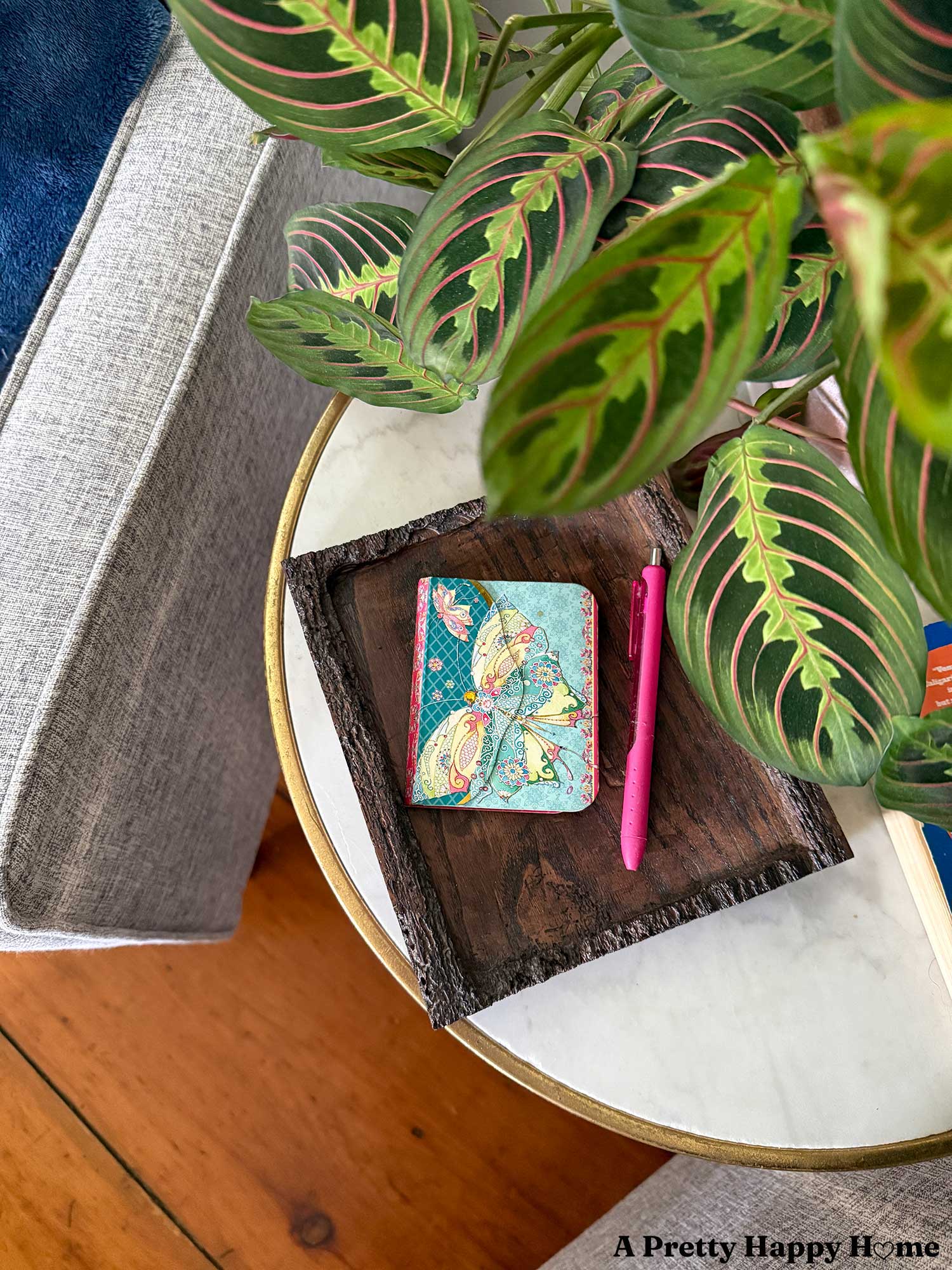 patterned notepad next to a pink pen sitting on a wood tray on a marble-topped round side table. There is a prayer plant leafing over the scene and the side table is flanked by two grey chairs.