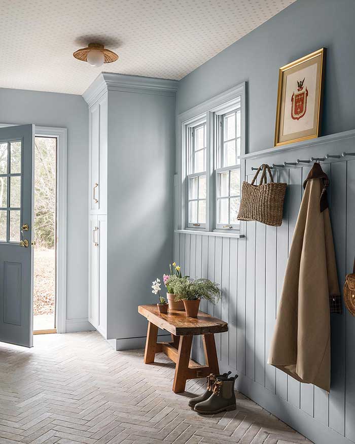light blue mudroom with wallpaper ceiling. Coat and bag hang from peg hooks and a wood bench holds plants.Design by Ann Lane Home and photography by Peak Visuals