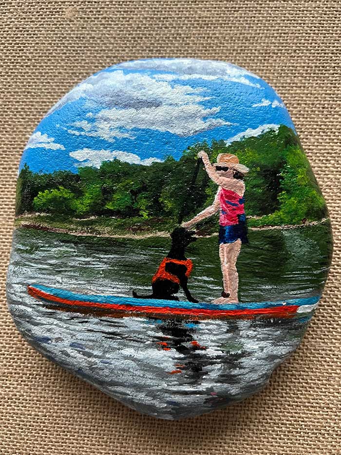 custom painted rock of a woman and a dog wearing a life jacket on a paddle board on a lake with green trees in the background