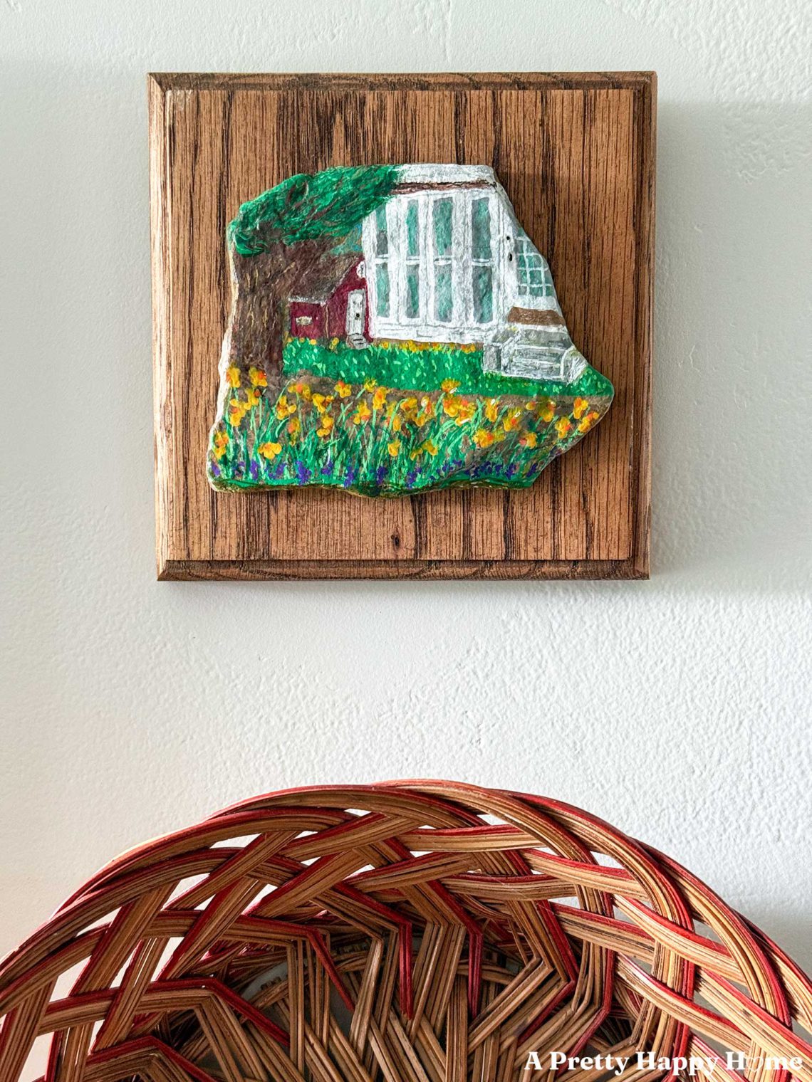 corner of a house and part of a red barn with daffodils in front of it painted on a small flat stone and mounted on a wood placard and hung above a basket on a wall.