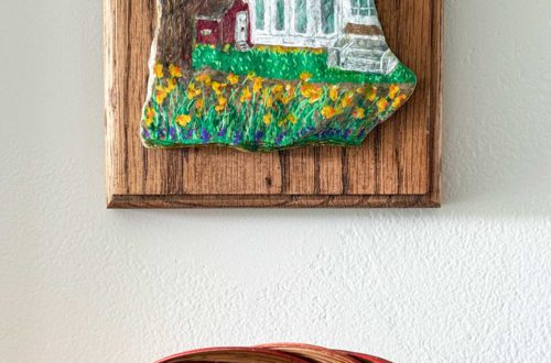 corner of a house and part of a red barn with daffodils in front of it painted on a small flat stone and mounted on a wood placard and hung above a basket on a wall.