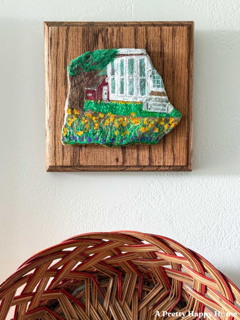 corner of a house and part of a red barn with daffodils in front of it painted on a small flat stone and mounted on a wood placard and hung above a basket on a wall.